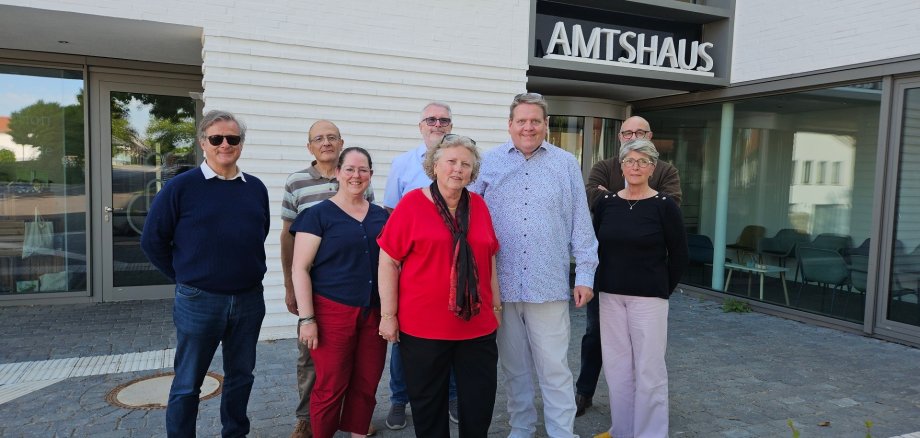 Sieben Menschen posieren vor einem weißen Gebäude, an dessen Fassade die Aufschrift Amtshaus zu lesen ist, für ein Gruppenfoto.