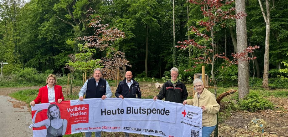 Mehrere Personen stehen in einem Waldstück vor frisch gepflanzten Bäumen und halten ein Banner hoch auf dem "Blutspende jetzt!" steht.