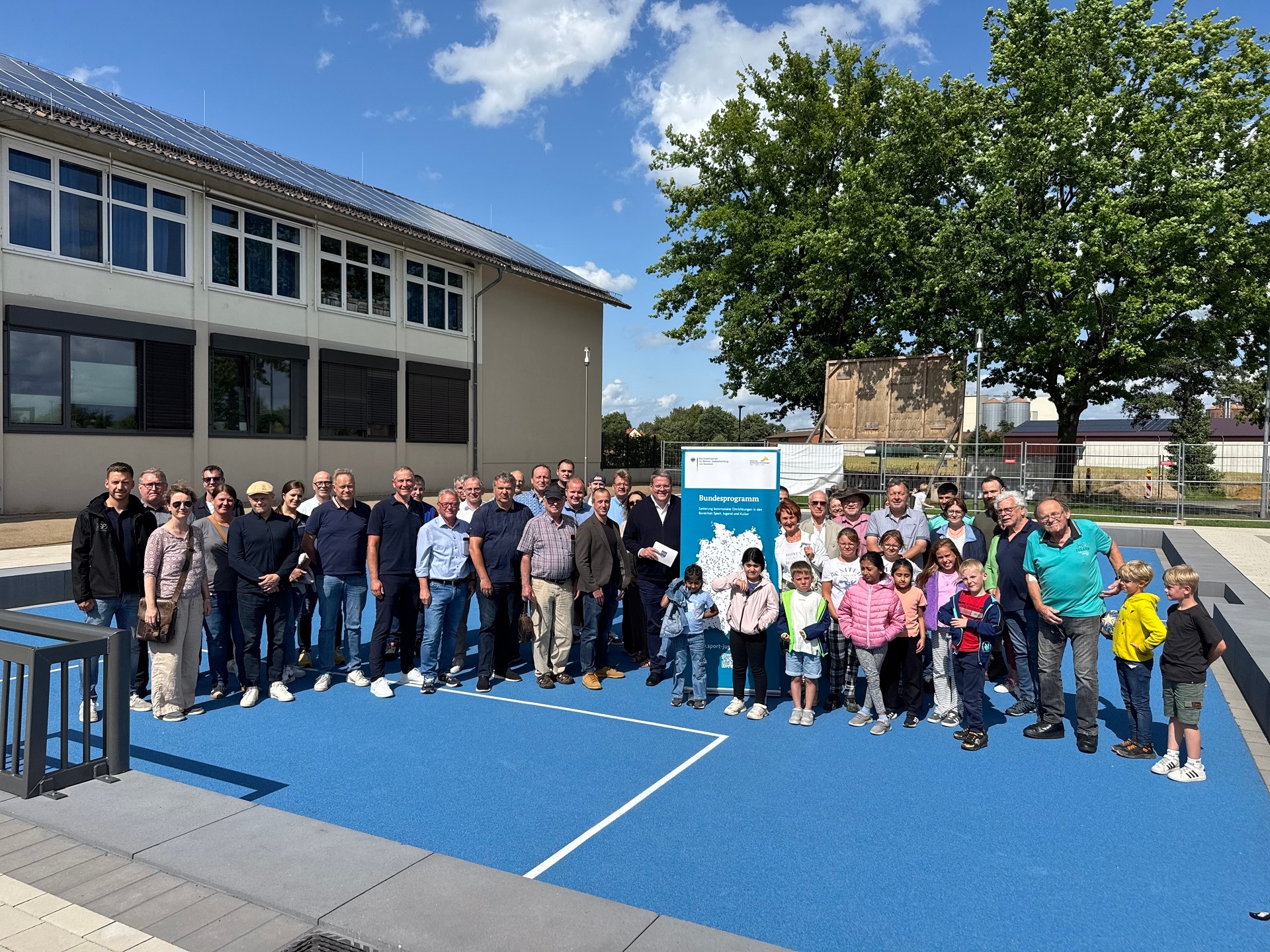 Gruppe von Erwachsenen und Kindern steht bei sonnigem Wetter auf einem neu gestalteten blauen Sportplatz vor einem Schulgebäude. Im Hintergrund ist ein Baum, ein Bauzaun sowie ein Aufsteller mit dem Logo des Bundesförderprogramms „Sanierung kommunaler Einrichtungen in den Bereichen Sport, Jugend und Kultur“ zu sehen. Alle Personen blicken zur Kamera, einige Kinder tragen bunte Jacken.