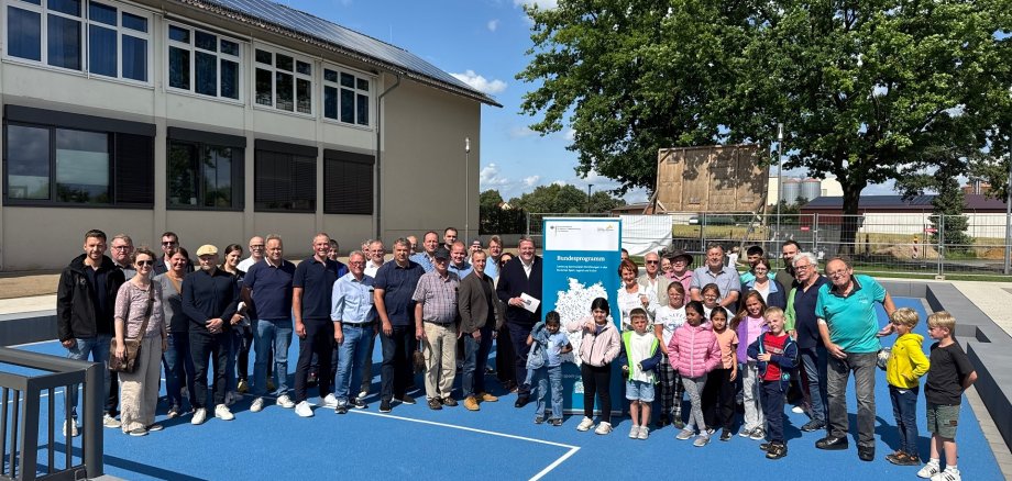 Gruppe von Erwachsenen und Kindern steht bei sonnigem Wetter auf einem neu gestalteten blauen Sportplatz vor einem Schulgebäude. Im Hintergrund ist ein Baum, ein Bauzaun sowie ein Aufsteller mit dem Logo des Bundesförderprogramms „Sanierung kommunaler Einrichtungen in den Bereichen Sport, Jugend und Kultur“ zu sehen. Alle Personen blicken zur Kamera, einige Kinder tragen bunte Jacken.