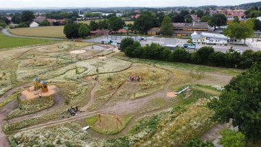 Naturspielplatz Wehdem Luftaufnahme eines weitläufigen Naturspielplatzes mit Wiesenflächen, Wegen, Spielgeräten und Gruppen von Kindern. Im Hintergrund sind Schulgebäude und ein Busbahnhof zu sehen.