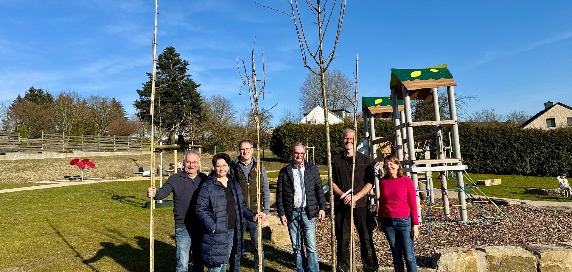 Sechs Personen haben sich vor einem Klettergerüst auf einem Spielplatz positioniert und präsentieren mehrere zum Einpflanzen bereite Bäume.
