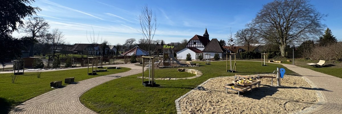 Mehrgenerationenpark Ein Spielplatzgelände, das auf einem leicht abschüssigen Gelände liegt und sich über mehrere Ebenen zieht. Es sind Bereiche mit Sand zu erkennen, Wege oder auch ein Klettergerüst und im Hintergrund eine historische Kapelle.