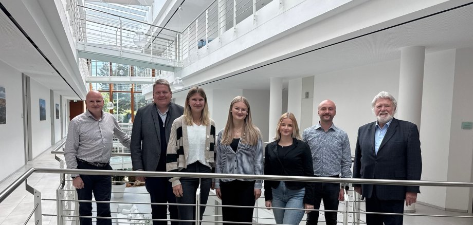 Insgesamt sieben Personen, vier Männer und drei junge Damen, haben sich in einem hellen Bürogebäude zum Gruppenfoto aufgestellt.