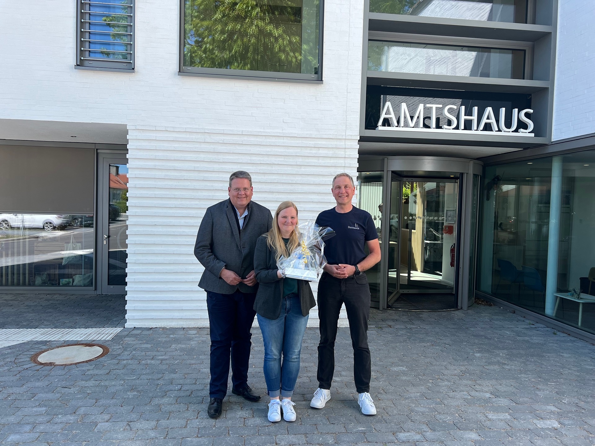 Zwei Männer stehen mit einer Frau, die ein Geschenk in den Händen hält, vor einem weißen Gebäude, auf dem der Schriftzug Amtshaus zu erkennen ist und lächeln in die Kamera.