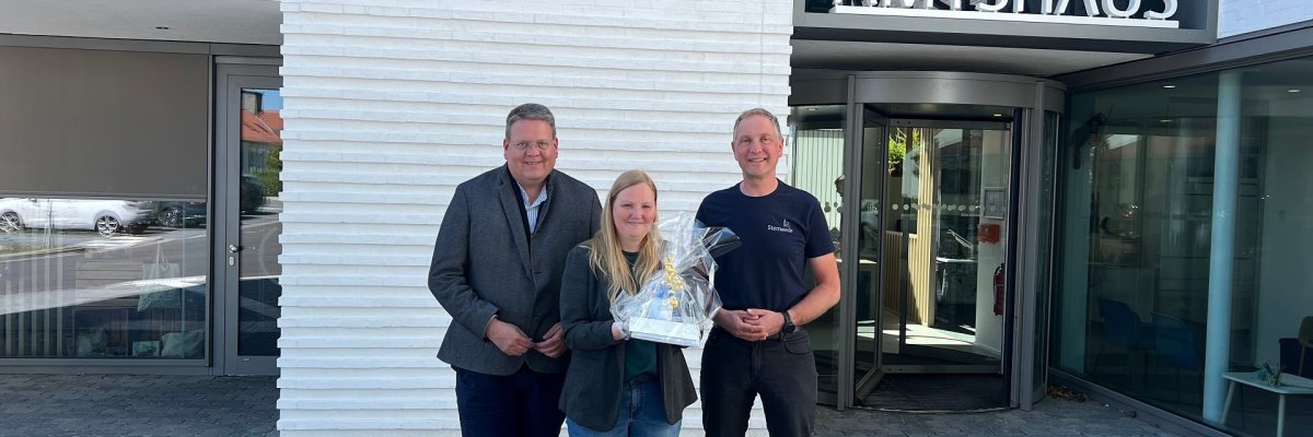 Zwei Männer stehen mit einer Frau, die ein Geschenk in den Händen hält, vor einem weißen Gebäude, auf dem der Schriftzug Amtshaus zu erkennen ist und lächeln in die Kamera.