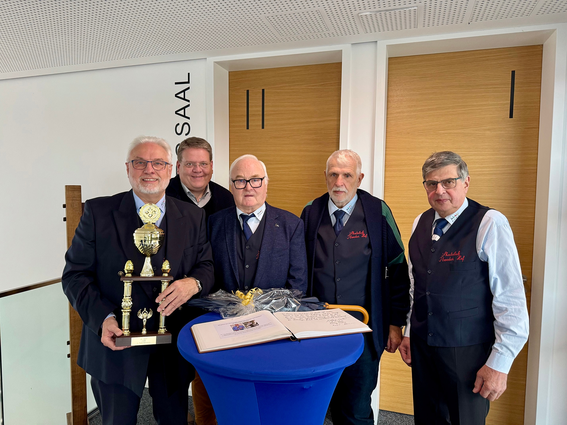 Fünf ältere Männer stehen gemeinsam in einem hellen Raum vor zwei Holztüren. Einer von ihnen hält einen großen goldenen Pokal. Auf einem blauen Stehtisch vor ihnen liegen ein aufgeschlagenes Gästebuch und ein Blumenstrauß. Drei der Männer tragen dunkle Westen mit gestickter Aufschrift „Skatclub Ilweder Hof“. Alle lächeln freundlich in die Kamera.