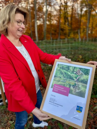 Klimaschutzpreis 2025 Eine Frau in einem roten Blazer, die einen Bilderrahmen hält, in dem eine Urkunde für den Klimaschutzpreis 2025 zu erkennen ist.