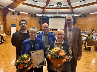 Dulli-Preis 20252 Fünf Personen stehen in einem großen Saal mit Holzwänden und Holzbalkendecke. Zwei Frauen in der vorderen Reihe halten Blumensträuße und eine gerahmte Urkunde mit der Aufschrift „Urkunde Dulli-Preis 2025“. Hinter ihnen stehen drei Männer. Im Hintergrund sitzen mehrere Personen an Tischen in einer Besprechungssituation. Die Atmosphäre wirkt feierlich, als ob eine Preisverleihung stattfindet.