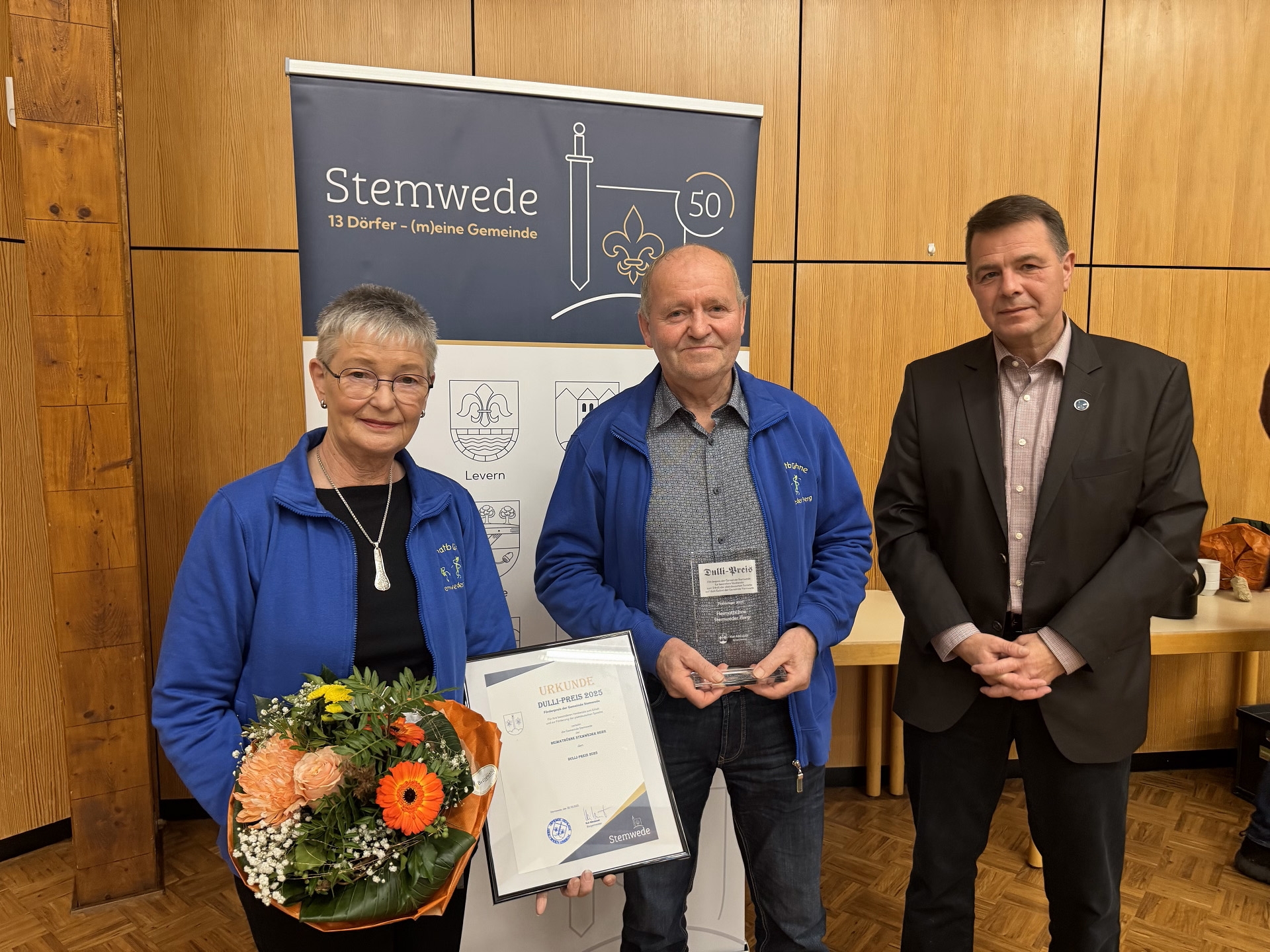 Drei Personen stehen in einem Raum mit Holzverkleidung. Links hält eine Frau einen Blumenstrauß und eine gerahmte Urkunde mit der Aufschrift „Dulli-Preis 2025“. Neben ihr steht ein Mann, der eine Glasplakette in den Händen hält. Beide tragen blaue Jacken mit Vereinslogo. Rechts steht ein Mann im dunklen Anzug. Hinter ihnen ist ein Roll-up mit der Aufschrift „Stemwede – 13 Dörfer (m)eine Gemeinde“ und grafischen Symbolen. Die Szene wirkt feierlich, als würde eine Auszeichnung überreicht.