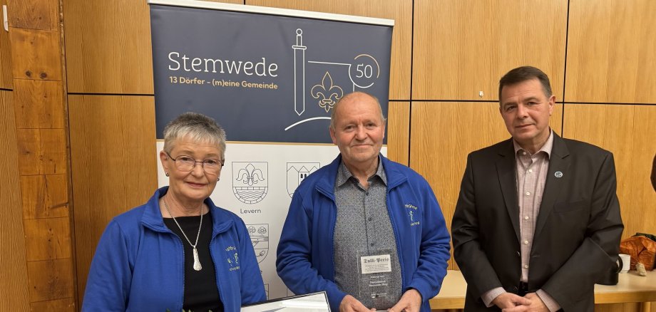 Dulli-Preis 2025 Drei Personen stehen in einem Raum mit Holzverkleidung. Links hält eine Frau einen Blumenstrauß und eine gerahmte Urkunde mit der Aufschrift „Dulli-Preis 2025“. Neben ihr steht ein Mann, der eine Glasplakette in den Händen hält. Beide tragen blaue Jacken mit Vereinslogo. Rechts steht ein Mann im dunklen Anzug. Hinter ihnen ist ein Roll-up mit der Aufschrift „Stemwede – 13 Dörfer (m)eine Gemeinde“ und grafischen Symbolen. Die Szene wirkt feierlich, als würde eine Auszeichnung überreicht.