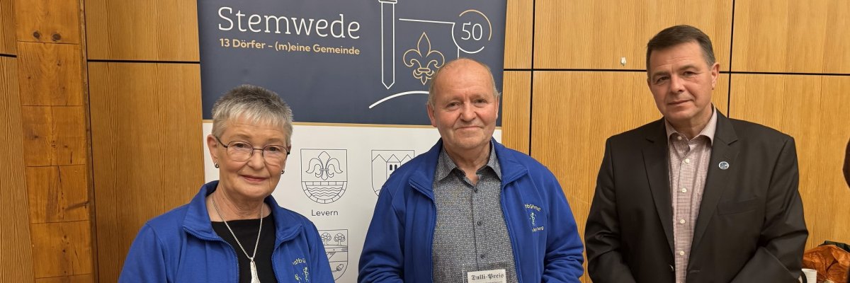 Dulli-Preis 2025 Drei Personen stehen in einem Raum mit Holzverkleidung. Links hält eine Frau einen Blumenstrauß und eine gerahmte Urkunde mit der Aufschrift „Dulli-Preis 2025“. Neben ihr steht ein Mann, der eine Glasplakette in den Händen hält. Beide tragen blaue Jacken mit Vereinslogo. Rechts steht ein Mann im dunklen Anzug. Hinter ihnen ist ein Roll-up mit der Aufschrift „Stemwede – 13 Dörfer (m)eine Gemeinde“ und grafischen Symbolen. Die Szene wirkt feierlich, als würde eine Auszeichnung überreicht.