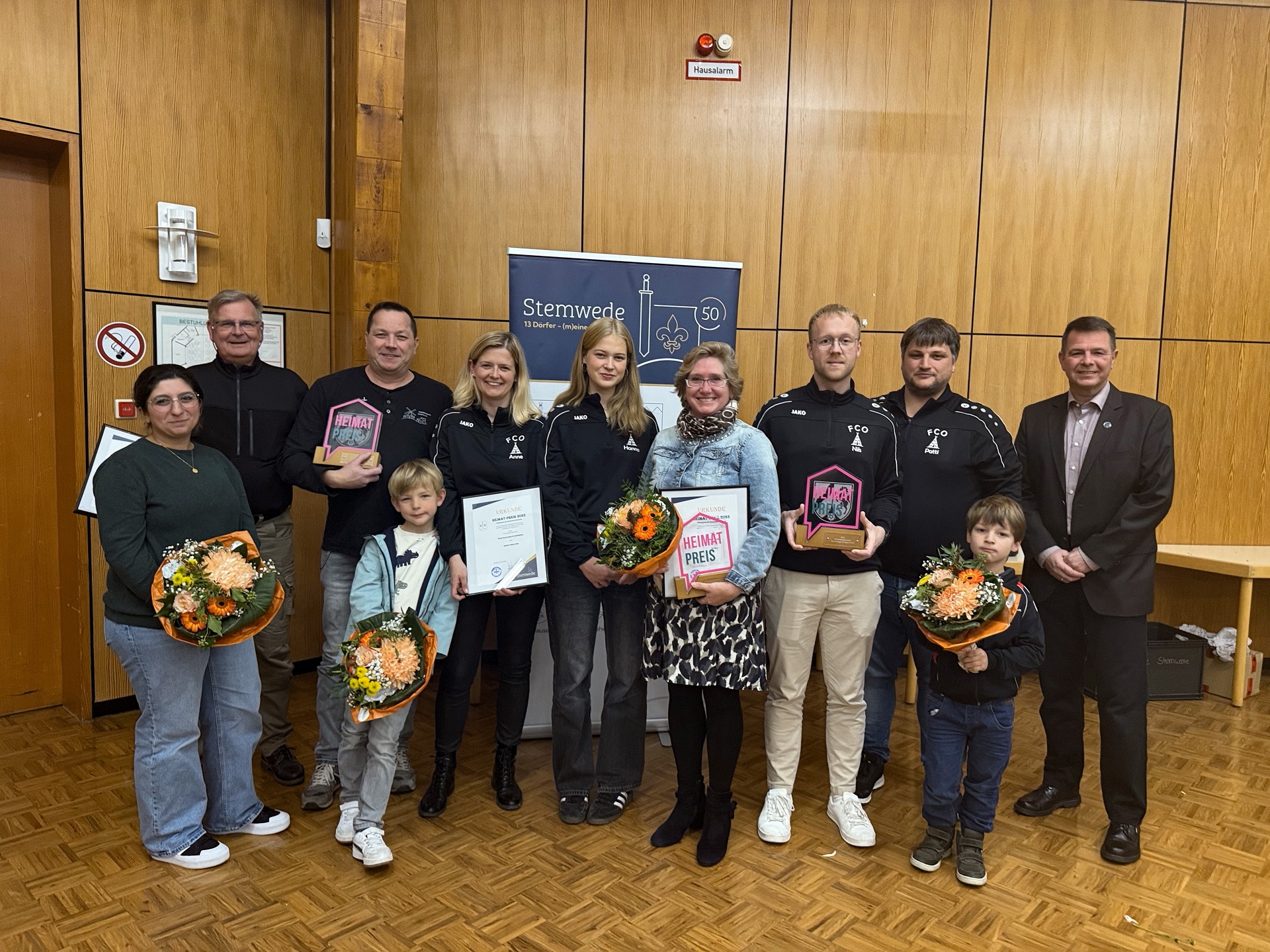 Eine Gruppe von elf Personen steht in einem Saal vor einer Holzvertäfelung. Mehrere Personen halten Urkunden, Glas-Trophäen mit der Aufschrift „Heimat-Preis“ und Blumensträuße in den Händen. Zwei Kinder stehen vorne. In der Mitte ist ein Banner mit dem Schriftzug „Stemwede“ zu sehen. Es handelt sich um eine Preisverleihung, bei der das Engagement von Bürgerinnen und Bürgern gewürdigt wird.