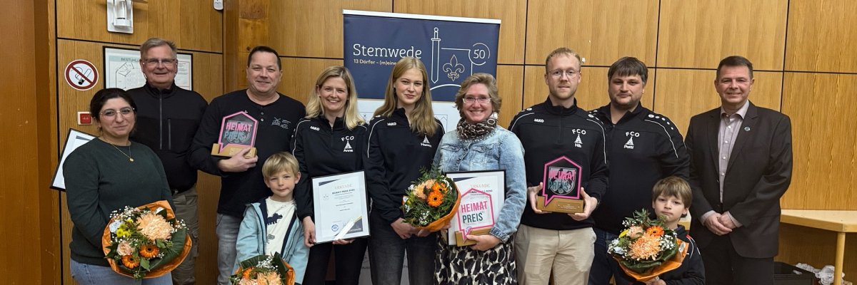 Eine Gruppe von elf Personen steht in einem Saal vor einer Holzvertäfelung. Mehrere Personen halten Urkunden, Glas-Trophäen mit der Aufschrift „Heimat-Preis“ und Blumensträuße in den Händen. Zwei Kinder stehen vorne. In der Mitte ist ein Banner mit dem Schriftzug „Stemwede“ zu sehen. Es handelt sich um eine Preisverleihung, bei der das Engagement von Bürgerinnen und Bürgern gewürdigt wird.