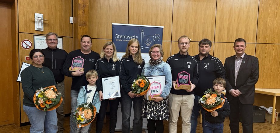 Eine Gruppe von elf Personen steht in einem Saal vor einer Holzvertäfelung. Mehrere Personen halten Urkunden, Glas-Trophäen mit der Aufschrift „Heimat-Preis“ und Blumensträuße in den Händen. Zwei Kinder stehen vorne. In der Mitte ist ein Banner mit dem Schriftzug „Stemwede“ zu sehen. Es handelt sich um eine Preisverleihung, bei der das Engagement von Bürgerinnen und Bürgern gewürdigt wird.