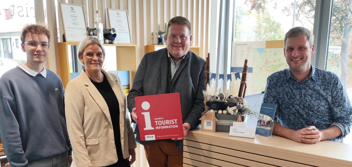 Vier Personen stehen in einem modernen Empfangsbereich einer Tourist-Information. Drei Personen stehen links neben dem Tresen, eine Person sitzt oder steht dahinter. Die Person in der Mitte hält ein rotes Schild mit einem weißen Informationssymbol und der Aufschrift „i – Certified Tourist Information“. Auf dem Tresen stehen kleine Souvenirartikel und Informationsmaterialien. Im Hintergrund sind Regale mit Dekoration und ein Fenster, durch das Bäume zu sehen sind. Die Atmosphäre ist freundlich und einladend.