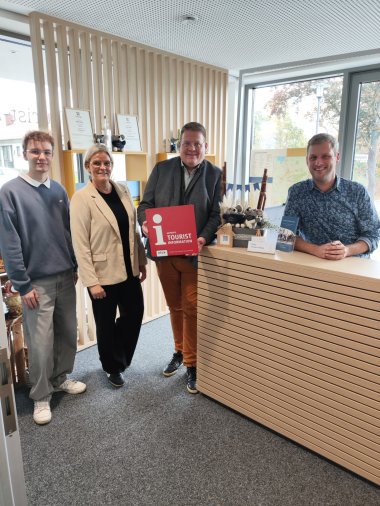 Tourist-i Vier Personen stehen in einem modernen Empfangsbereich einer Tourist-Information. Drei Personen stehen links neben dem Tresen, eine Person sitzt oder steht dahinter. Die Person in der Mitte hält ein rotes Schild mit einem weißen Informationssymbol und der Aufschrift „i – Certified Tourist Information“. Auf dem Tresen stehen kleine Souvenirartikel und Informationsmaterialien. Im Hintergrund sind Regale mit Dekoration und ein Fenster, durch das Bäume zu sehen sind. Die Atmosphäre ist freundlich und einladend.