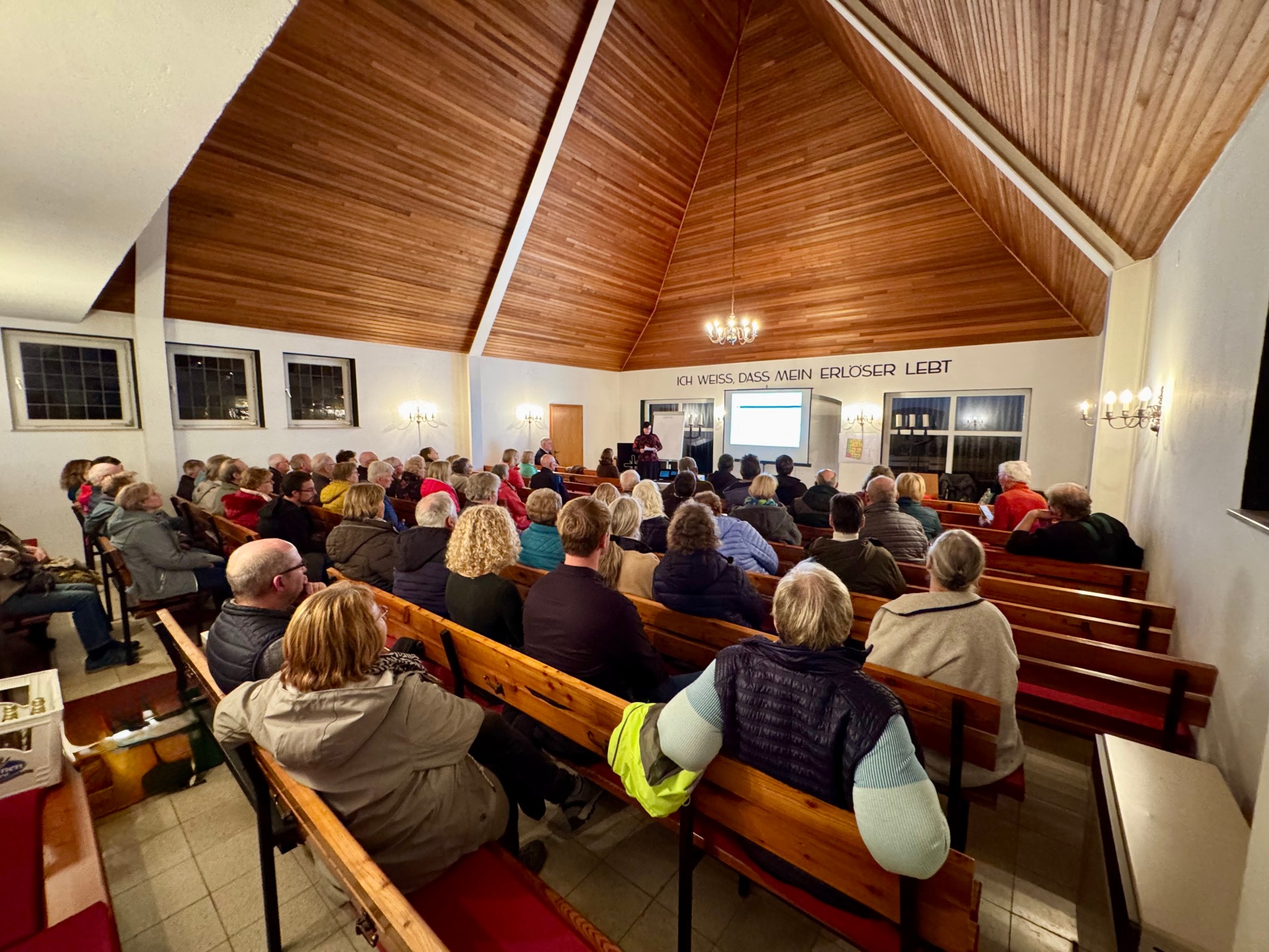 In einem kirchlichen Gemeinderaum mit Holzdecke sitzen zahlreiche Menschen auf Holzbänken und verfolgen eine Präsentation. Vorne steht eine Person an einem Rednerpult, hinter ihr ist eine Leinwand mit einer Projektion zu sehen. An der Wand über dem Altarraum steht der Schriftzug „Ich weiß, dass mein Erlöser lebt“. Die Veranstaltung wirkt gut besucht und findet in einer ruhigen, warm beleuchteten Atmosphäre statt.
