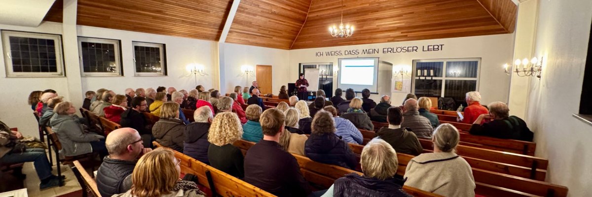 Infoveranstaltung Drohne In einem kirchlichen Gemeinderaum mit Holzdecke sitzen zahlreiche Menschen auf Holzbänken und verfolgen eine Präsentation. Vorne steht eine Person an einem Rednerpult, hinter ihr ist eine Leinwand mit einer Projektion zu sehen. An der Wand über dem Altarraum steht der Schriftzug „Ich weiß, dass mein Erlöser lebt“. Die Veranstaltung wirkt gut besucht und findet in einer ruhigen, warm beleuchteten Atmosphäre statt.