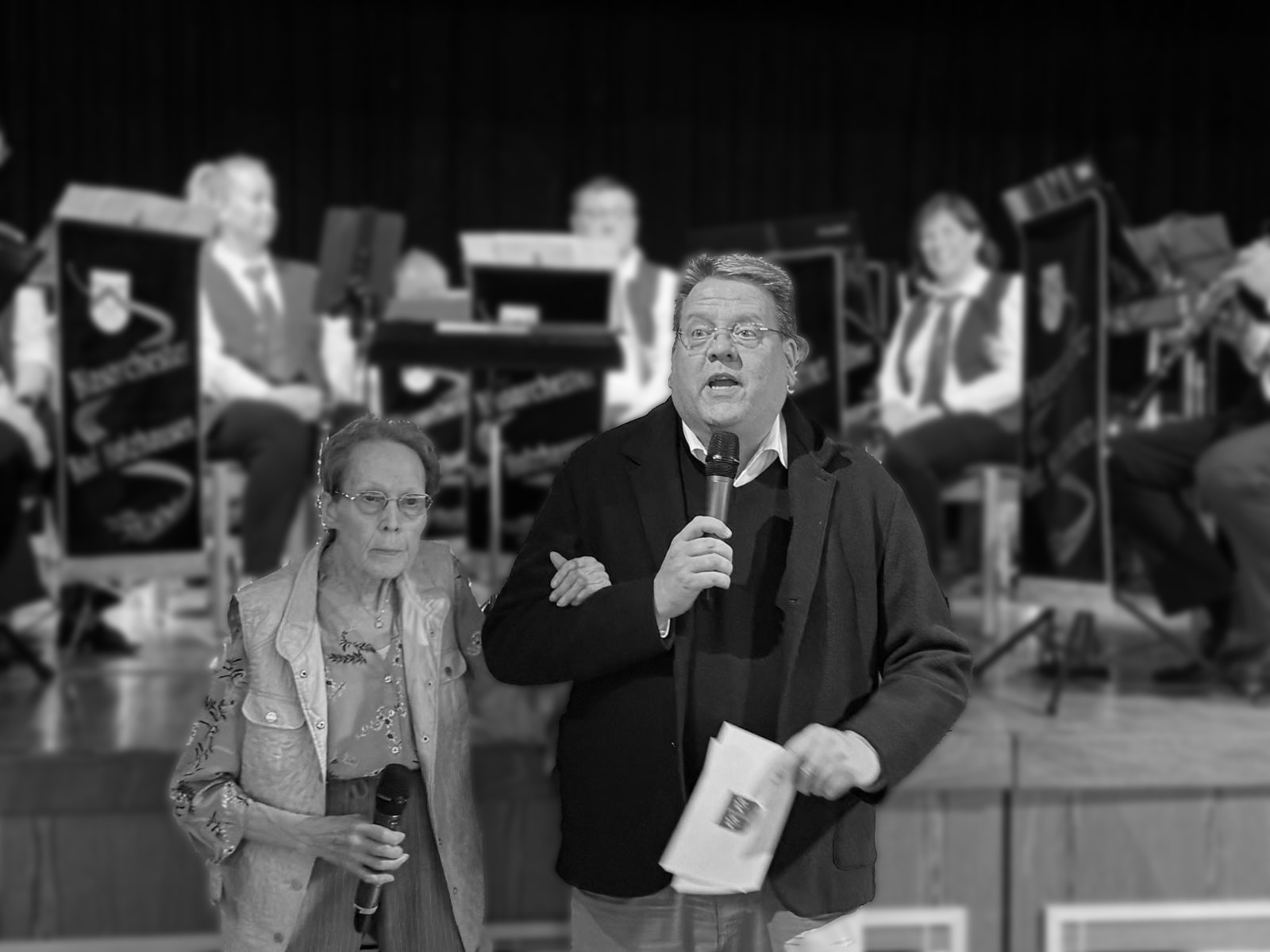 Eine schwarz-weiß-Aufnahme zweier Personen, die gerade scheinbar ein Publikum begrüßen. Im Hintergrund ist verschwommen eine Blaskapelle zu erkennen. Da es eine schwarz-weiß-Aufnahme ist, scheint es sich um einen Nachruf zu handeln.