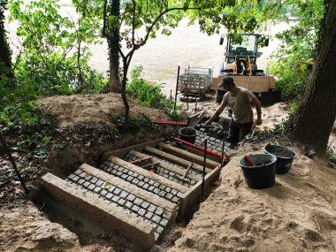 Treppe in Westrup Eine im Bau befindliche Außentreppe wird von einer Person in Arbeitskleidung saniert. Die Stufen bestehen aus neu verlegten, quadratischen Natursteinpflastersteinen und werden von seitlichen Betonwangen eingefasst. Um die Treppe herum liegen Eimer, Werkzeuge und Baumaterial. Hinter der Baustelle steht ein kleiner Radlader sowie Materialwagen auf einer offenen, sandigen Fläche. Die Umgebung ist bewaldet und dicht begrünt.