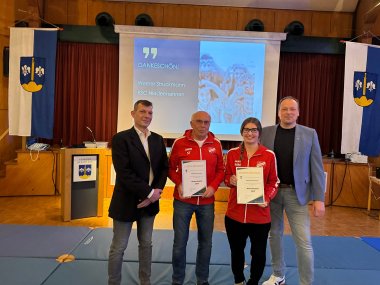 Eine Frau und ein Mann in roten Pullis stehen vor einer Bühne. Auf einer Leinwand steht "Ehrenamtspreis, Werner Struckmann, RSC Niedermehnen". Links und rechts von den beiden Personen stehen zwei Männer in zivil.
