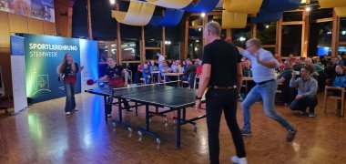 Vier Personen spielen Tischtennis in einem voll besetzten und dekoriertem Festsaal.