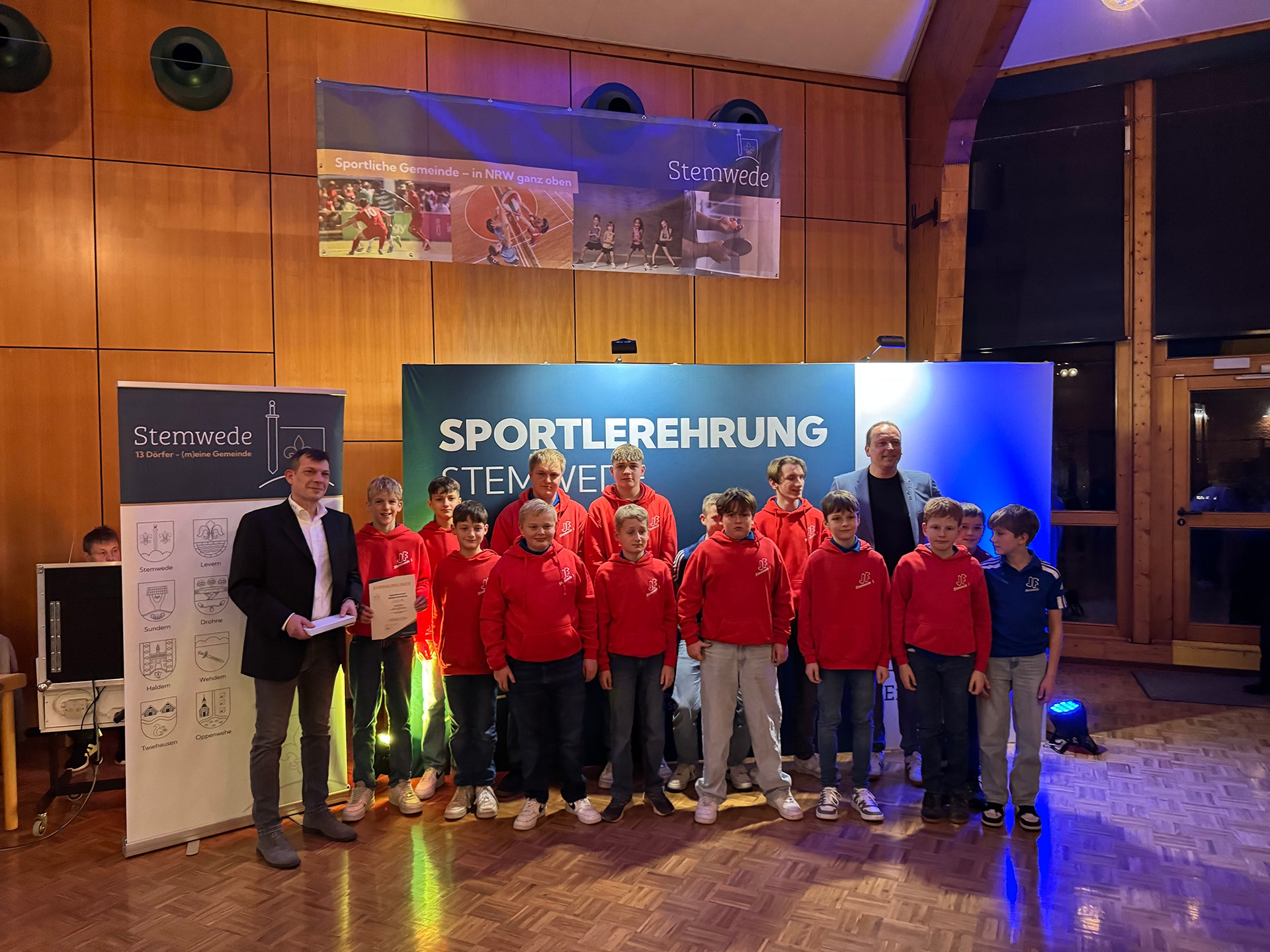 Viele Personen in roten Vereinspullis stehen für ein Gruppenfoto vor einer blau-weißen Bühnenwand mit der Aufschrift „Sportlerehrung Stemwede“. Links ist ein Banner der Gemeinde Stemwede zu sehen, oben hängen Sportfotos als Dekoration. Die Szene ist warm ausgeleuchtet und wirkt feierlich.