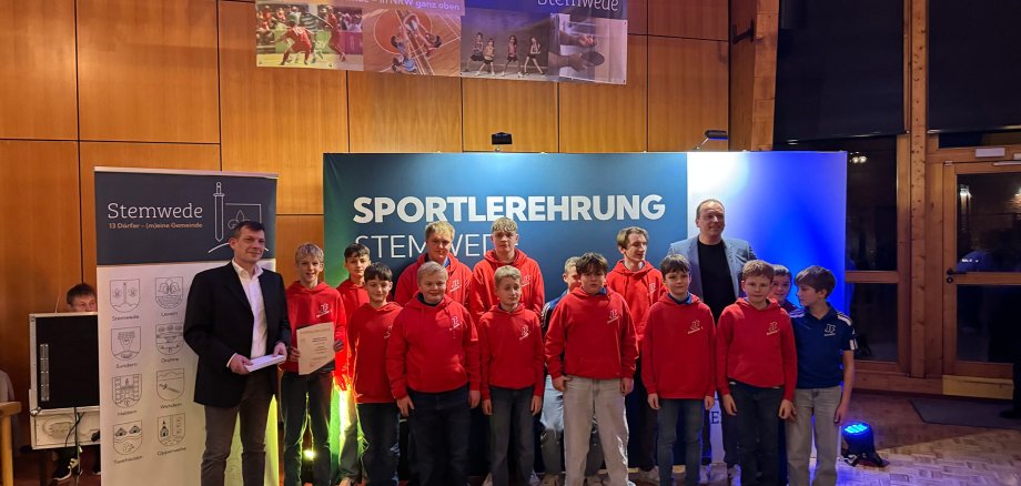 Viele Personen in roten Vereinspullis stehen für ein Gruppenfoto vor einer blau-weißen Bühnenwand mit der Aufschrift „Sportlerehrung Stemwede“. Links ist ein Banner der Gemeinde Stemwede zu sehen, oben hängen Sportfotos als Dekoration. Die Szene ist warm ausgeleuchtet und wirkt feierlich.