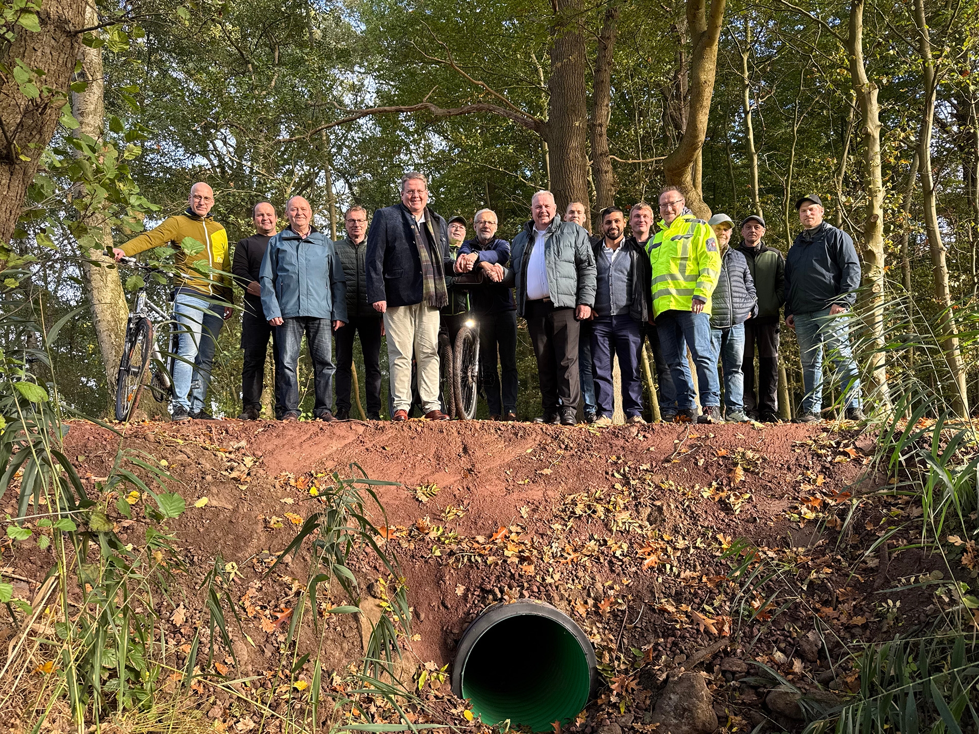 Eine Gruppe von etwa 15 Personen steht auf einem frisch angelegten Erdwall in einem Waldgebiet, vor ihnen ein grünes Rohr als Durchlass. Die Menschen posieren für ein Foto, einige tragen Outdoor- oder Arbeitskleidung.