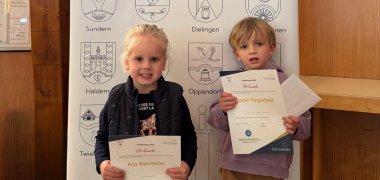 Ehrung STADTRADELN 2025 Zwei kleine Kinder stehen vor einem Roll-up-Banner mit der Aufschrift „Stemwede – 13 Dörfer (m)eine Gemeinde“. Beide halten Urkunden in den Händen und lächeln in die Kamera. Das Banner zeigt oben das Wappen von Stemwede und unten die Wappen der 13 Ortsteile. Der Hintergrund besteht aus einer hölzernen Wandverkleidung.