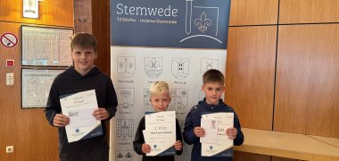 Ehrung STADTRADELN 2025 Drei Jungs stehen vor einem Roll-up-Banner mit der Aufschrift „Stemwede – 13 Dörfer (m)eine Gemeinde“. Beide halten Urkunden in den Händen und lächeln in die Kamera. Das Banner zeigt oben das Wappen von Stemwede und unten die Wappen der 13 Ortsteile. Der Hintergrund besteht aus einer hölzernen Wandverkleidung.