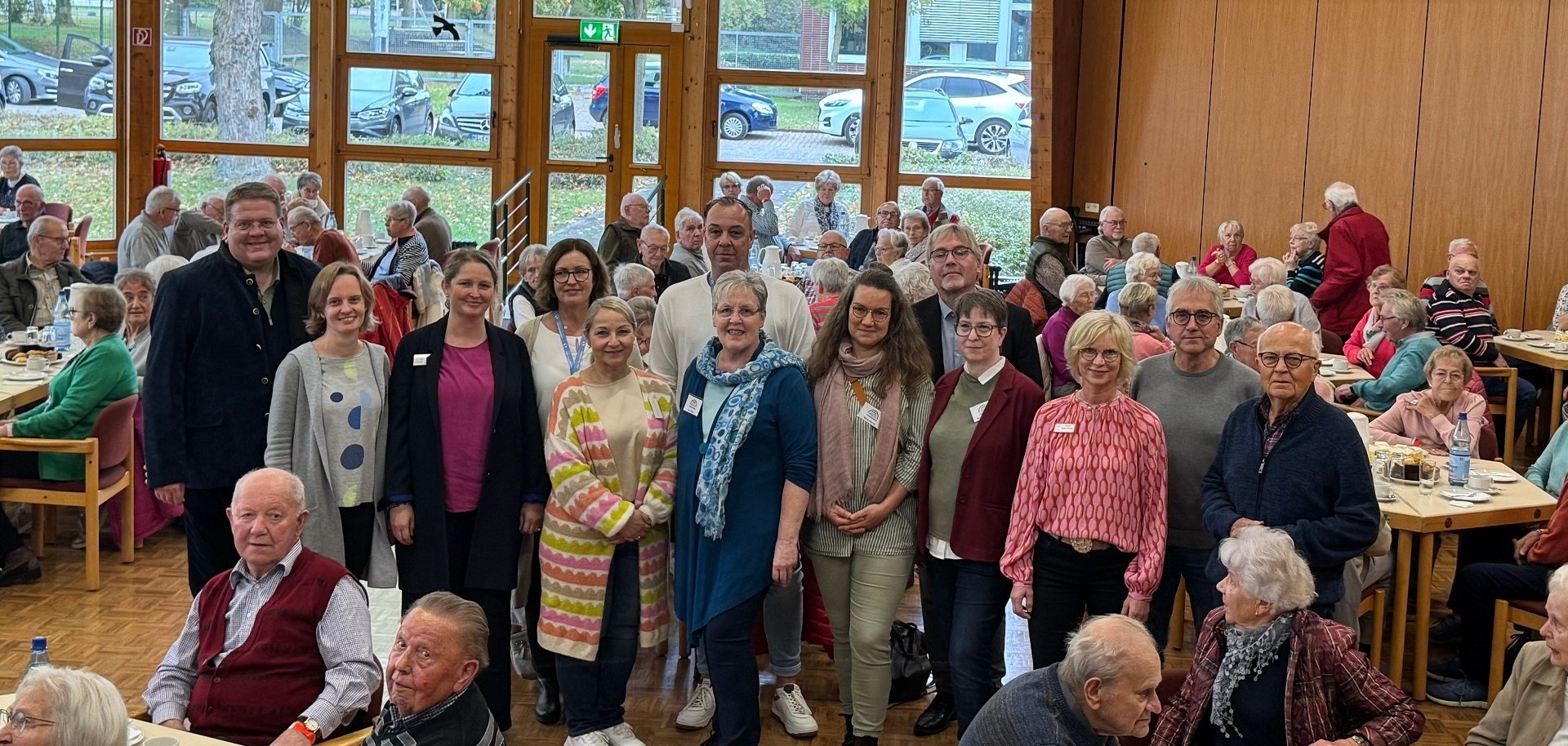 Eine große Gruppe von älteren Menschen sitzt in einem hellen Saal mit großen Fenstern an Tischen, auf denen Kaffee und Kuchen stehen. Im Vordergrund steht eine Gruppe von etwa 15 Personen, die in die Kamera lächeln – vermutlich Organisatorinnen und Organisatoren der Veranstaltung. Im Hintergrund sind viele Seniorinnen und Senioren, die sich unterhalten. Durch die Fenster sieht man herbstliche Bäume und geparkte Autos. Die Stimmung wirkt freundlich und gemeinschaftlich.