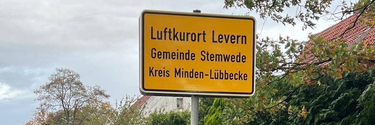 Ortseingangsschild Zwei Männer stehen lächelnd neben einem gelben Ortseingangsschild an einer Landstraße. Auf dem Schild steht: „Luftkurort Levern, Gemeinde Stemwede, Kreis Minden-Lübbecke“. Im Hintergrund sind eine Hecke, ein Haus mit rotem Dach und herbstliche Bäume zu sehen. Der Himmel ist bewölkt, der Straßenrand ist feucht.