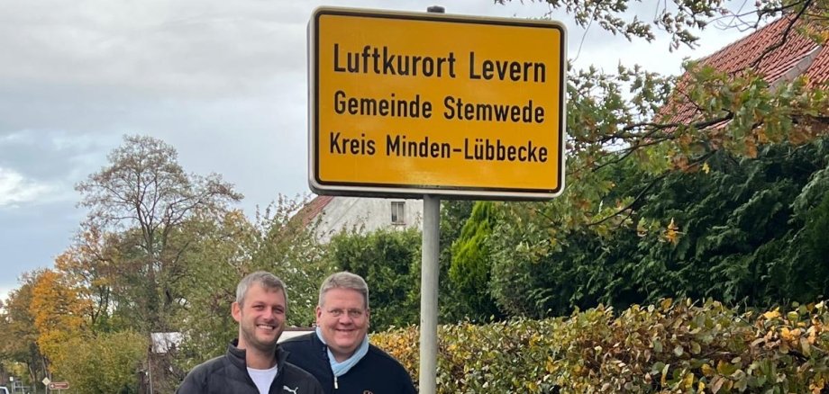 Ortseingangsschild Zwei Männer stehen lächelnd neben einem gelben Ortseingangsschild an einer Landstraße. Auf dem Schild steht: „Luftkurort Levern, Gemeinde Stemwede, Kreis Minden-Lübbecke“. Im Hintergrund sind eine Hecke, ein Haus mit rotem Dach und herbstliche Bäume zu sehen. Der Himmel ist bewölkt, der Straßenrand ist feucht.