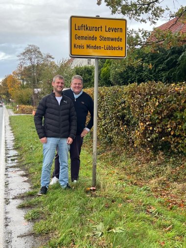 Ortseingangsschild Zwei Männer stehen lächelnd neben einem gelben Ortseingangsschild an einer Landstraße. Auf dem Schild steht: „Luftkurort Levern, Gemeinde Stemwede, Kreis Minden-Lübbecke“. Im Hintergrund sind eine Hecke, ein Haus mit rotem Dach und herbstliche Bäume zu sehen. Der Himmel ist bewölkt, der Straßenrand ist feucht.