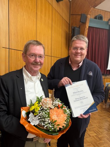 Zwei Männer stehen nebeneinander. Der linke hält einen Blumenstrauße in der Hand, der rechte eine Urkunde.