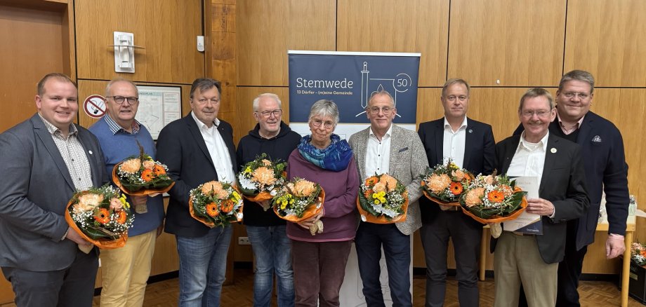 Eine Gruppe von neun Personen steht lächelnd in einem Raum vor einer Holzvertäfelung. Alle halten Blumensträuße in den Händen. In der Mitte hängt ein Banner mit der Aufschrift „Stemwede – 13 Dörfer, eine Gemeinde“. Es scheint sich um eine Ehrung oder Auszeichnung zu handeln.