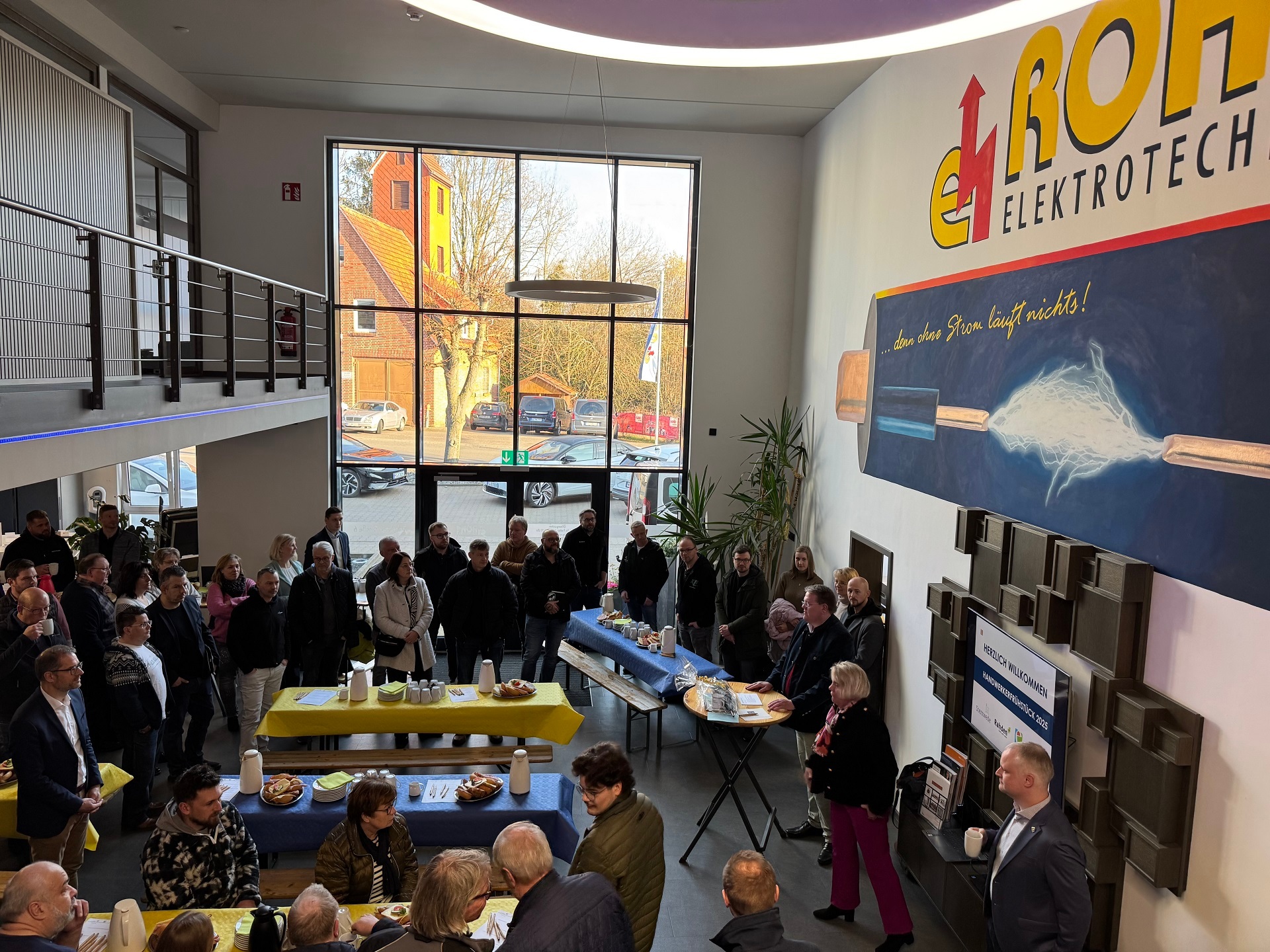 Zahlreiche Personen im Foyer eines Unternehmens. Die Aufnahme wurde von der Galerie auf die Personen im Erdgeschoss gemacht. Die Personen scheinen stehen und sitzen im Foyer und scheinen einem Redner zuzuhören.