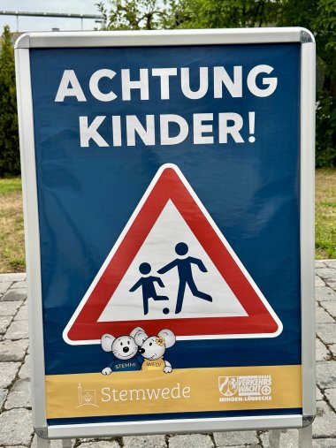 Achtung Kinder Mobiles Hinweisschild mit der Aufschrift „ACHTUNG KINDER!“ und einem Verkehrszeichen, das auf spielende Kinder hinweist. Darunter sind die Stemweder Maskottchen „Stemmi“ und „Wedi“ abgebildet. Im unteren Bereich stehen die Logos und Schriftzüge der Gemeinde Stemwede und der Kreis-Verkehrswacht Minden-Lübbecke.