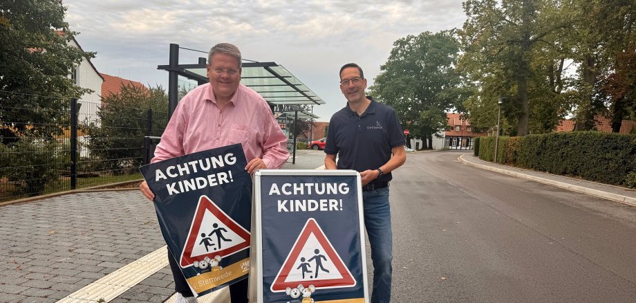 Achtung Kinder Zwei Männer stehen an einer Straße in Stemwede und präsentieren gemeinsam zwei Hinweisschilder mit der Aufschrift „ACHTUNG KINDER!“ und einem Verkehrszeichen, das auf Kinder im Straßenverkehr aufmerksam macht. Im Hintergrund sind eine Bushaltestelle, Wohnhäuser und Bäume zu sehen.