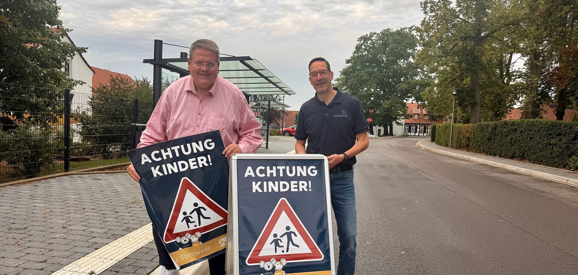 Zwei Männer stehen an einer Straße in Stemwede und präsentieren gemeinsam zwei Hinweisschilder mit der Aufschrift „ACHTUNG KINDER!“ und einem Verkehrszeichen, das auf Kinder im Straßenverkehr aufmerksam macht. Im Hintergrund sind eine Bushaltestelle, Wohnhäuser und Bäume zu sehen.
