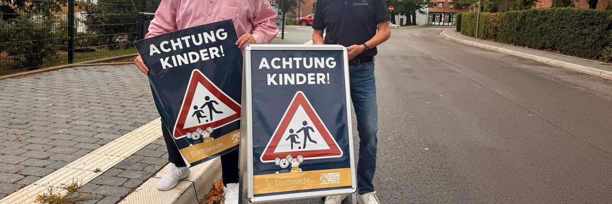 Achtung Kinder Zwei Männer stehen an einer Straße in Stemwede und präsentieren gemeinsam zwei Hinweisschilder mit der Aufschrift „ACHTUNG KINDER!“ und einem Verkehrszeichen, das auf Kinder im Straßenverkehr aufmerksam macht. Im Hintergrund sind eine Bushaltestelle, Wohnhäuser und Bäume zu sehen.