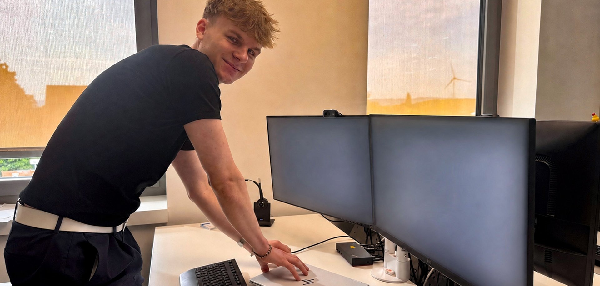 Ein junger Auszubildender steht lächelnd an einem modernen Büroarbeitsplatz und schließt ein silbernes Notebook an eine Dockingstation an. Vor ihm befinden sich zwei große Monitore, eine Tastatur und eine Maus auf einem hellen Schreibtisch. Im Hintergrund sorgen große Fenster mit heruntergelassenen Rollos für Tageslicht; draußen ist unter anderem ein Windrad zu erkennen. Die Monitore zeigen keine lesbaren Inhalte.