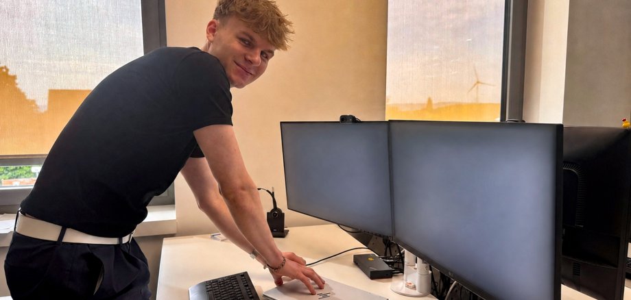 IT-Azubi 2026 Ein junger Auszubildender steht lächelnd an einem modernen Büroarbeitsplatz und schließt ein silbernes Notebook an eine Dockingstation an. Vor ihm befinden sich zwei große Monitore, eine Tastatur und eine Maus auf einem hellen Schreibtisch. Im Hintergrund sorgen große Fenster mit heruntergelassenen Rollos für Tageslicht; draußen ist unter anderem ein Windrad zu erkennen. Die Monitore zeigen keine lesbaren Inhalte.