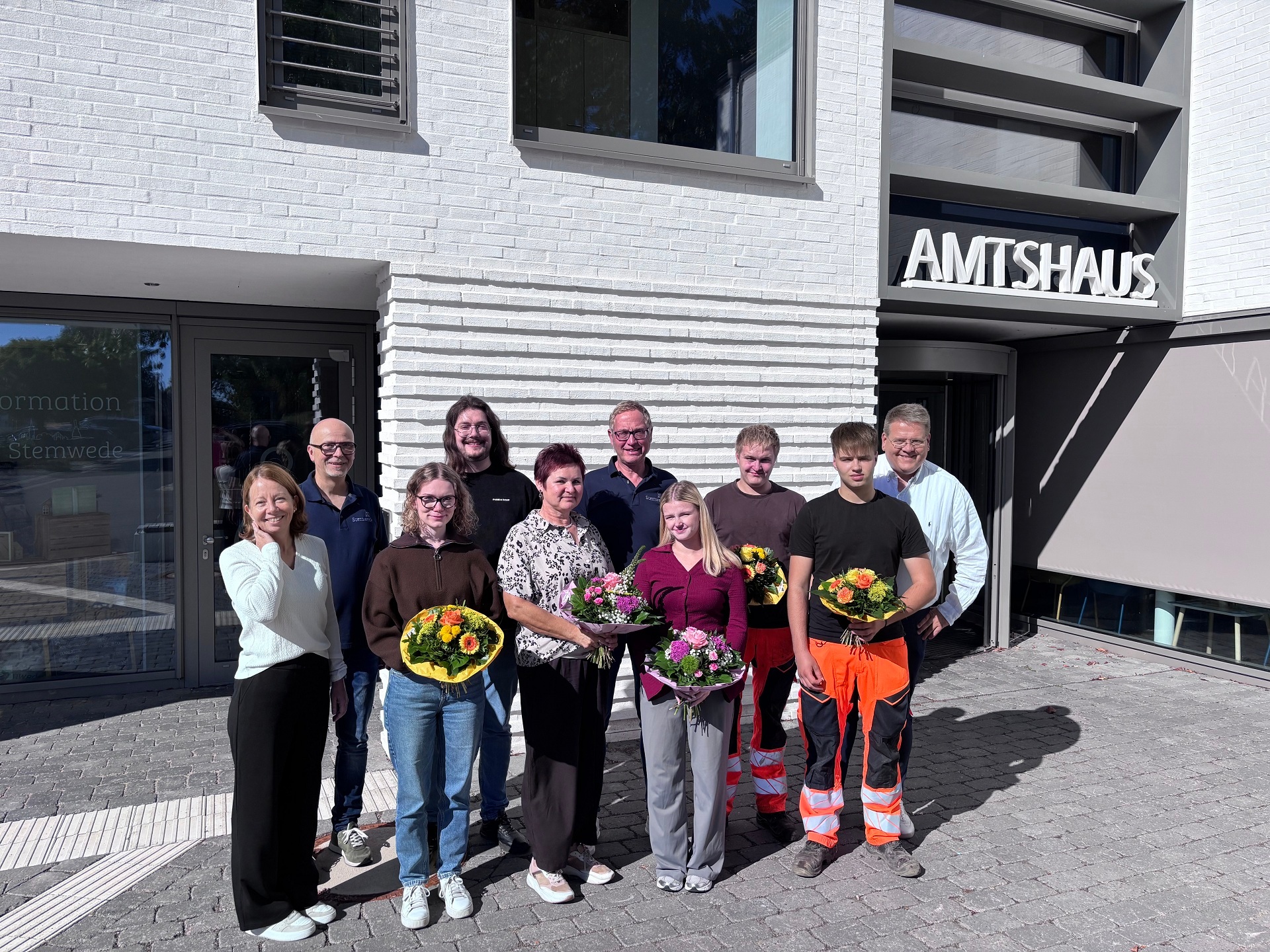 Gruppenfoto vor dem Stemweder Amtshaus: Mehrere Mitarbeiterinnen und Mitarbeiter stehen bei Sonnenschein vor dem Eingang. Vier Personen in der ersten Reihe halten bunte Blumensträuße.