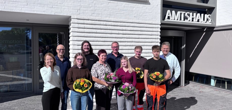 Absolventen 2026 Gruppenfoto vor dem Stemweder Amtshaus: Mehrere Mitarbeiterinnen und Mitarbeiter stehen bei Sonnenschein vor dem Eingang. Vier Personen in der ersten Reihe halten bunte Blumensträuße.