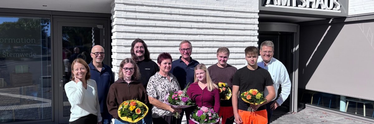 Absolventen 2026 Gruppenfoto vor dem Stemweder Amtshaus: Mehrere Mitarbeiterinnen und Mitarbeiter stehen bei Sonnenschein vor dem Eingang. Vier Personen in der ersten Reihe halten bunte Blumensträuße.