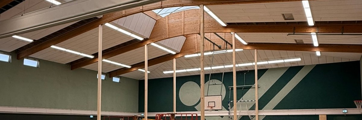 Blick in eine Sporthalle während der Sanierungsarbeiten. Auf dem Hallenboden liegen Holzplatten und Baumaterialien, mehrere Hubarbeitsbühnen stehen in der Halle. Im Hintergrund sind Basketballkörbe, Handballtore und die markante Hallenwand zu sehen. An der Decke sind große Holzbinder, Oberlichter und neue LED-Leuchten erkennbar.