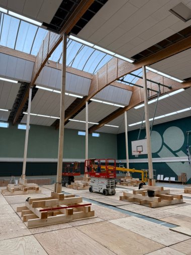 Blick in eine Sporthalle während der Sanierungsarbeiten. Auf dem Hallenboden sind großflächige Holzplatten verlegt, darauf stehen mehrere Hubarbeitsbühnen und Holzbauteile. Deutlich sichtbar sind die hölzernen Dachträger, die lichtdurchlässigen Oberlichter und die neuen Deckenverkleidungen. Im Hintergrund befinden sich ein Basketballkorb und eine große grafische Gestaltung an der Hallenwand.